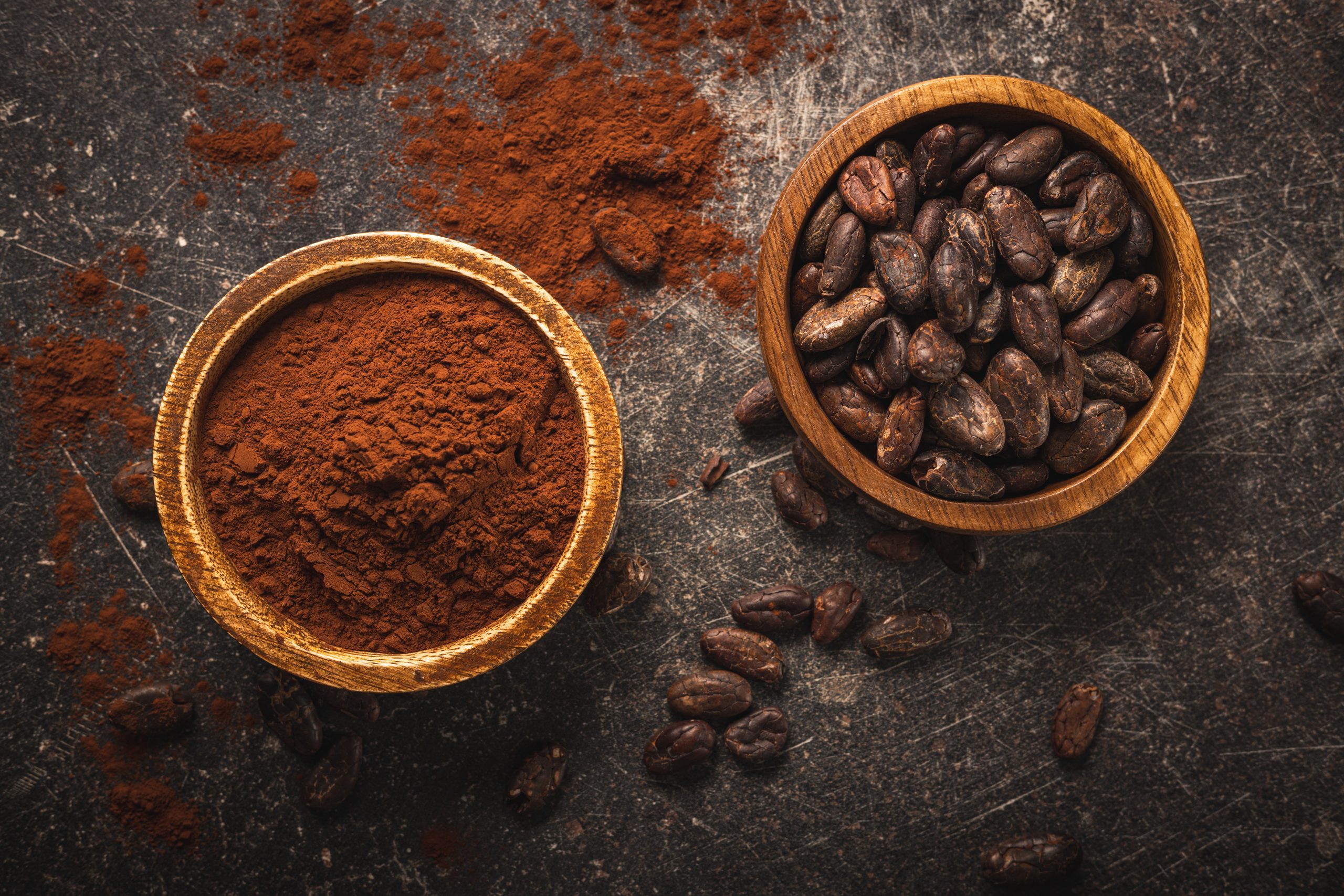 Cocoa beans and cocoa powder in wooden bowls on black table.